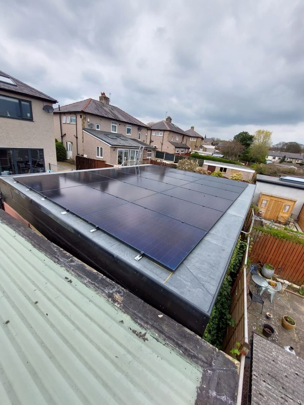 Black solar panels tilted slightly on the flat rubber roof of an extension, with suburban houses behind
