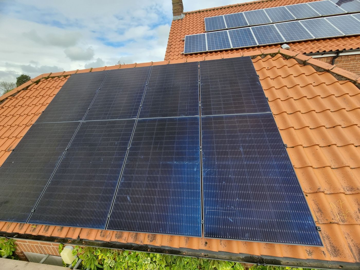 Close-up of new all-black solar panels on a clay pantile roof, with an older blue solar array higher up